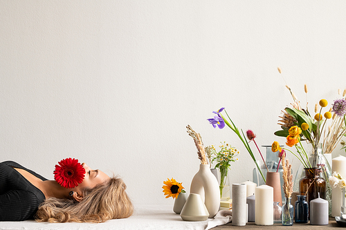 Young serene woman with red herbera in her mouth lying by variety of fresh and dried flowers and wildflowers in vases and group of candles