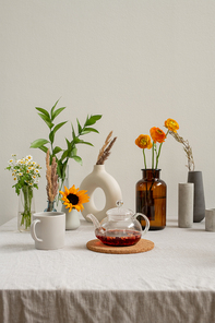 Teapot with fresh herbal tea and mug on background of various fresh flowers in vases standing on table against white wall in the kitchen