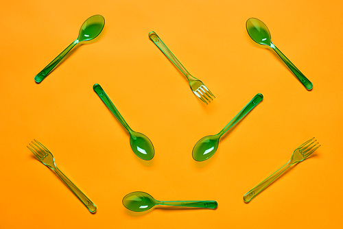 Horizontal flat lay conceptual shot of green plastic spoons and forks on bright orange background