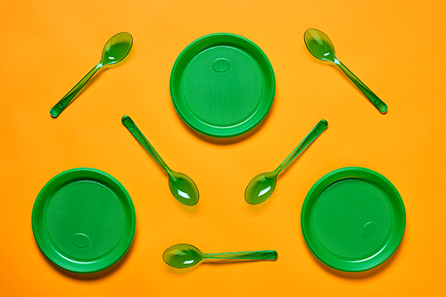 Horizontal flat lay conceptual shot of green plastic spoons and plates on bright orange background