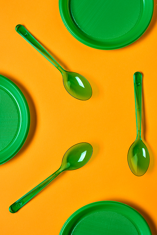 Vertical flat lay conceptual pattern shot of green plastic spoons and plates on bright orange background