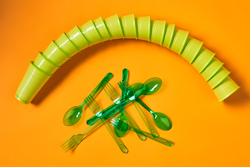 Horizontal flat lay shot of disposable green plastic cups set, spoons and forks on bright orange background