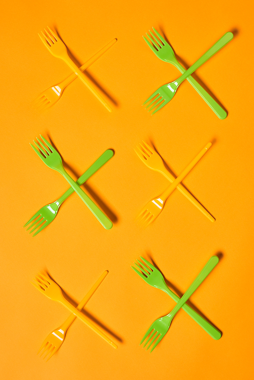Vertical conceptual flat lay shot of X marks made of disposable light green and orange plastic forks on orange background