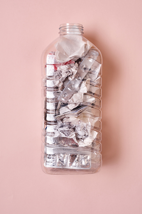 Transparent plastic bottle full of various garbage on pale pink background, vertical from above flat lay shot
