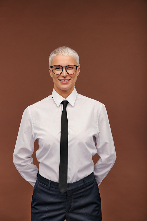 Cheerful stylish masculine woman in formalwear and eyeglasses looking at you with toothy smile while standing in front of camera in isolation
