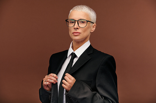 Young elegant blond woman in black suit, tie and eyeglasses looking at you with confident expression while standing in front of camera