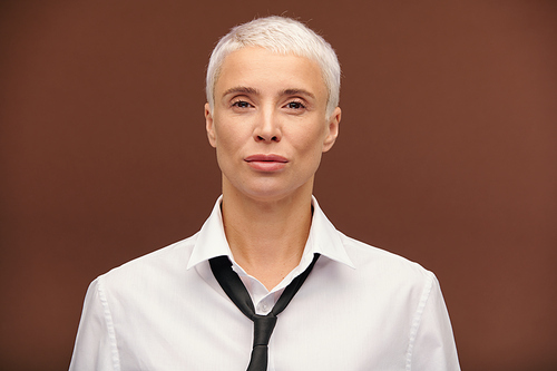 Serious mature blond businesswoman in white shirt and black tie standing in front of camera and looking at you against brown background