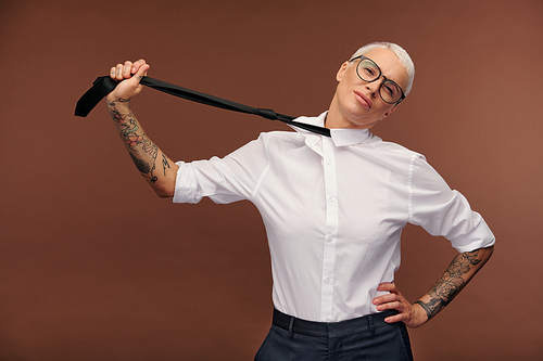 Mature masculine blond businesswoman with arms covered by tattoos stretching black necktie while keeping left hand on waist