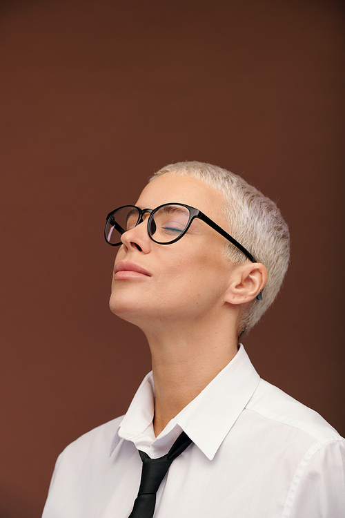 Contemporary young serene woman in eyeglasses, white shirt and black tie keeping her eyes closed and head slightly backwards in isolation