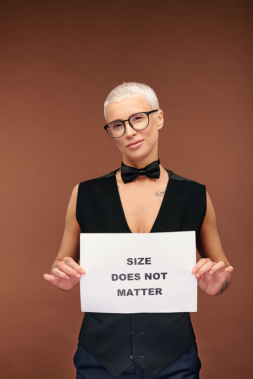 Young eccentric blond female with black bowtie and waistcoat on nude body holding paper poster saying that size does not matter