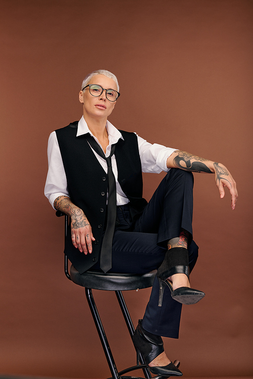 Young masculine elegant female with short blond hair and arms and body covered by tattoos sitting on chair in front of camera in isolation