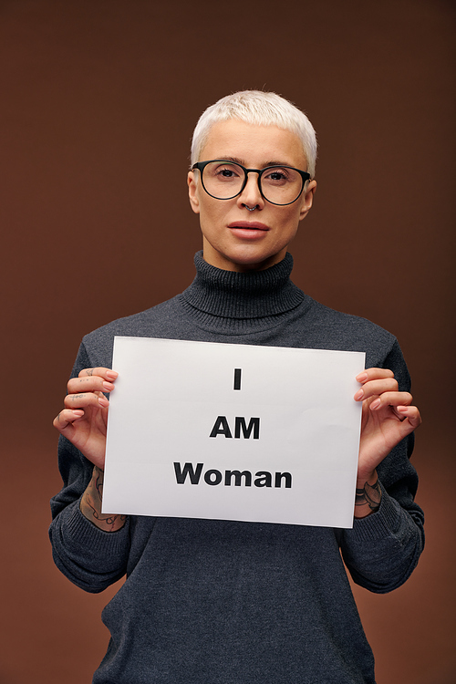Young elegant blond female in dark grey pullover and eyeglasses holding paper saying that she is a woman while standing in front of camera