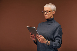Serious blond elegant businesswoman in dark grey pullover and eyeglasses using mobile gadget in front of camera while surfing in the net