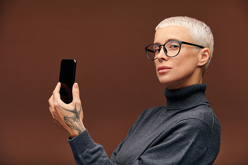Young serious blond businesswoman in pullover and eyeglasses holding smartphone in front of herself while looking at you in isolation