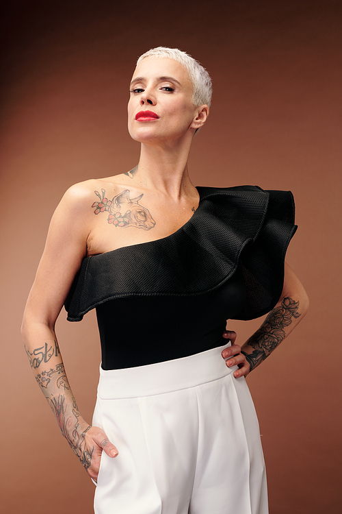 Elegant woman in black tanktop and white trousers with tattoos on arms and chest keeping her head slightly turned to the right in isolation