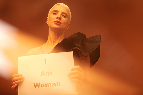 Blond elegant woman in black tanktop holding paper saying that she is woman while standing in front of camera on lit brown background