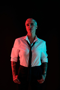 Silhouette of young elegant manlike female with short hair wearing white shirt, black tie and pants standing in front of camera in darkness