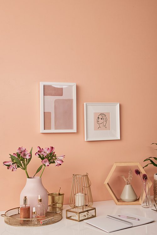 Workplace of interior designer by pink wall with two pictures in frames by table with flowers in vases, beauty care products, candles and other stuff