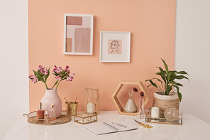 Workplace of interior designer by pink wall with two pictures in frames by table with flowers, beauty care products, candles and open copybook