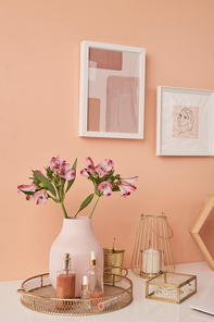 Two pictures in frames on pink wall by table with flowers in vase, beauty care products, candle, pearl necklace, golden bucket and other stuff