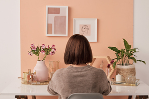 Back view of brunette female interior designer sitting by table in front of pink wall with two pictures in frames and carrying out her work