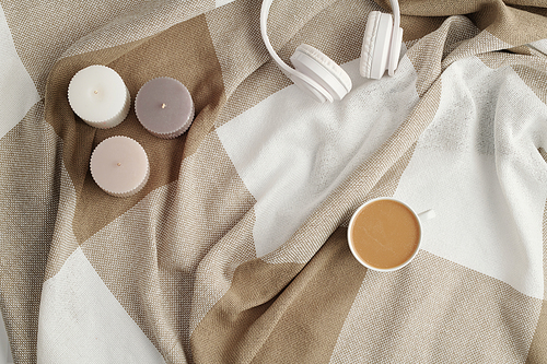 Flat lay of three small aromatic candles, white headphones and cup of fresh cappuccino on checkered linen or cotton plaid