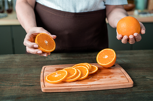 Hands of contemporary housewife in apron standing by kitchen table and holding fresh oranges over wooden board with juicy slices