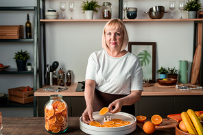 Blond middle aged housewife in t-shirt and apron looking at you while standing by kitchen table and putting orange slices on fruit dryer
