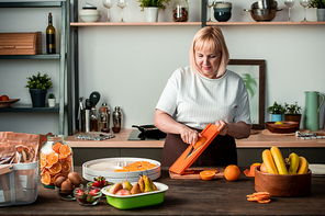 Blond pretty housewife standing by kitchen table and cutting fresh oranges among bowls with kiwi, strawberries, pears and bananas