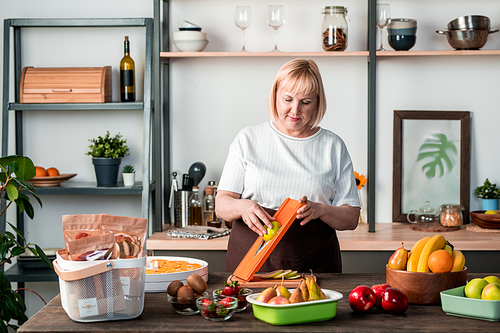 Blond female in casualwear standing by kitchen table with variety of fresh fruits and using cutter while preparing homemade dry pears