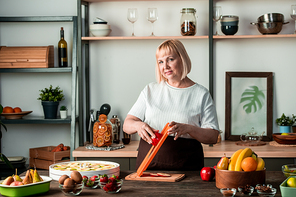 Blond female in casualwear standing by kitchen table with variety of fresh fruits and using cutter while preparing homemade dry pears