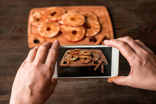 Hands of housewife with smartphone taking photo of slices of fresh apple sprinkled with ground cinnamon on wooden board and table