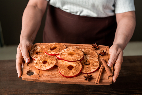 Hands of mature female holding chopping board with pile of fresh apple slices sprinkled with ground cinnamon over kitchen table