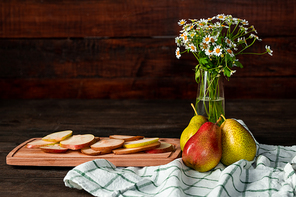 Still life composition with vase of wildflowers, linen kitchen towel, ripe garden pears and chopping board with slices of fresh fruit