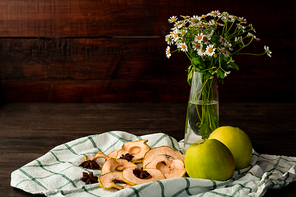 Fresh granny smith apples, vase with garden flowers, dry fruit and star anise on checkered kitchen towel on dark wooden table against wall