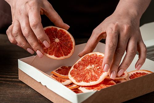 Hands of contemporary middle aged housewife putting homemade dried orange or grapefruit slices into small square carton box