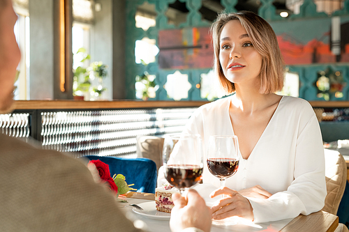 Pretty young elegant female with glass of red wine looking at her boyfriend while both sitting by table in restaurant during lunch