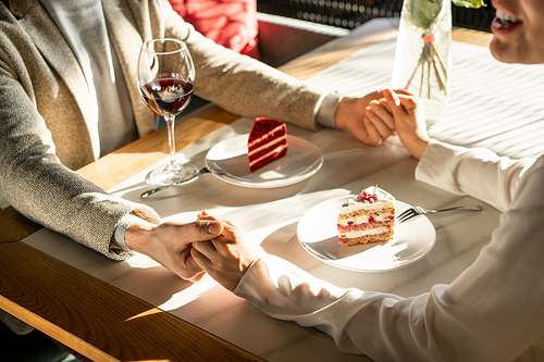 Two young romantic dates holding by hands over served table while having tasty cakes and red wine after lunch in classy restaurant