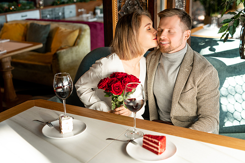 Young happy woman with luxurious bouquet of red roses kissing her boyfriend on cheek while both sitting by table in restaurant