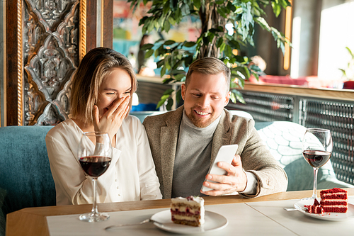 Horizontal shot of young man and woman sitting together at cafe table watching something funny on smartphone and laughing