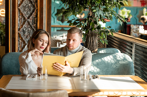 Young romantic couple looking through menu in luxurious restaurant and discussing what to choose while sitting on couch by table