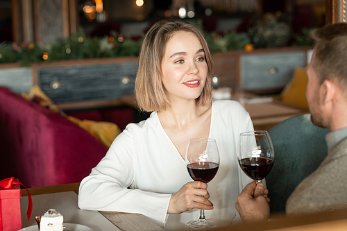 Horizontal portrait of beautiful woman in love sitting in front of man in modern restaurant holding glassess with wine chatting with him