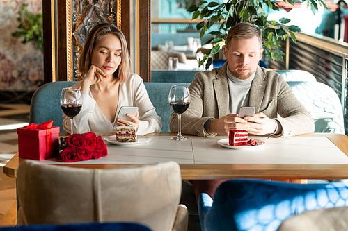 Young adult Caucasian man and woman sitting together in modern restaurant surfing Internet on their smartphones instead of talking to each other