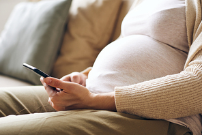 Close-up of unrecognizable pregnant woman in comfortable clothes sitting on sofa and using smartphone