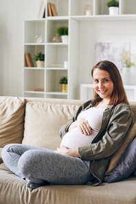 happy young pregnant female in casualwear sitting among pillows on couch in front of camera in home