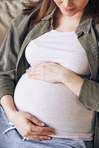 Close-up of young pregnant woman in casual clothing embracing belly while thinking about baby in her womb