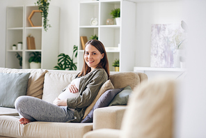 young cheerful pregnant woman with toothy smiling looking at you while resting on couch with cushions in home