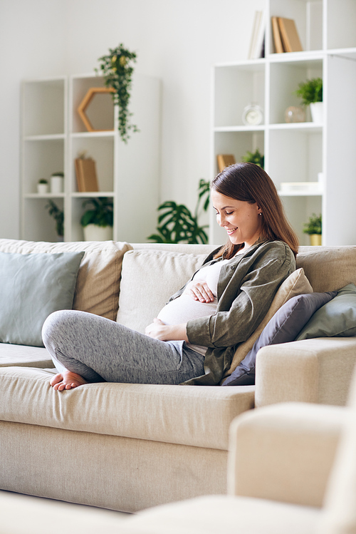 Happy young pregnant woman in casual outfit sitting with crossed legs on sofa in living room and talking to baby in her womb