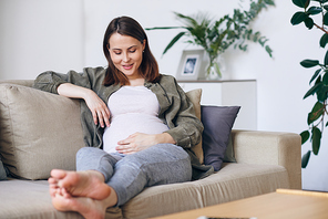 Young barefoot female in casualwear looking at her pregnant belly while relaxing on couch among cushions in living-room