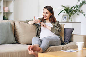 Smiling beautiful young pregnant woman in casual shirt sitting on sofa and using smartphone while reading internet articles about baby-raising
